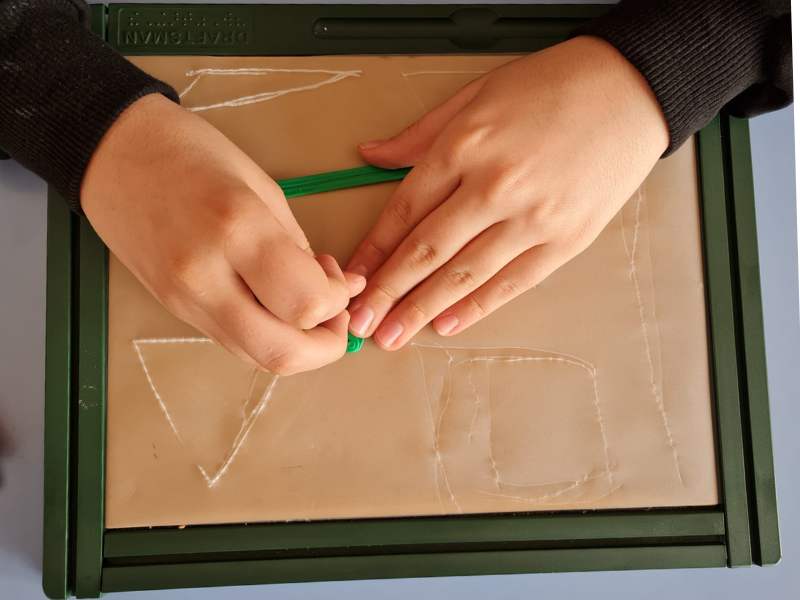 Fotoğrafta dokunsal çizim tabletine üçgen çizen bir çocuğun elleri görünüyor.